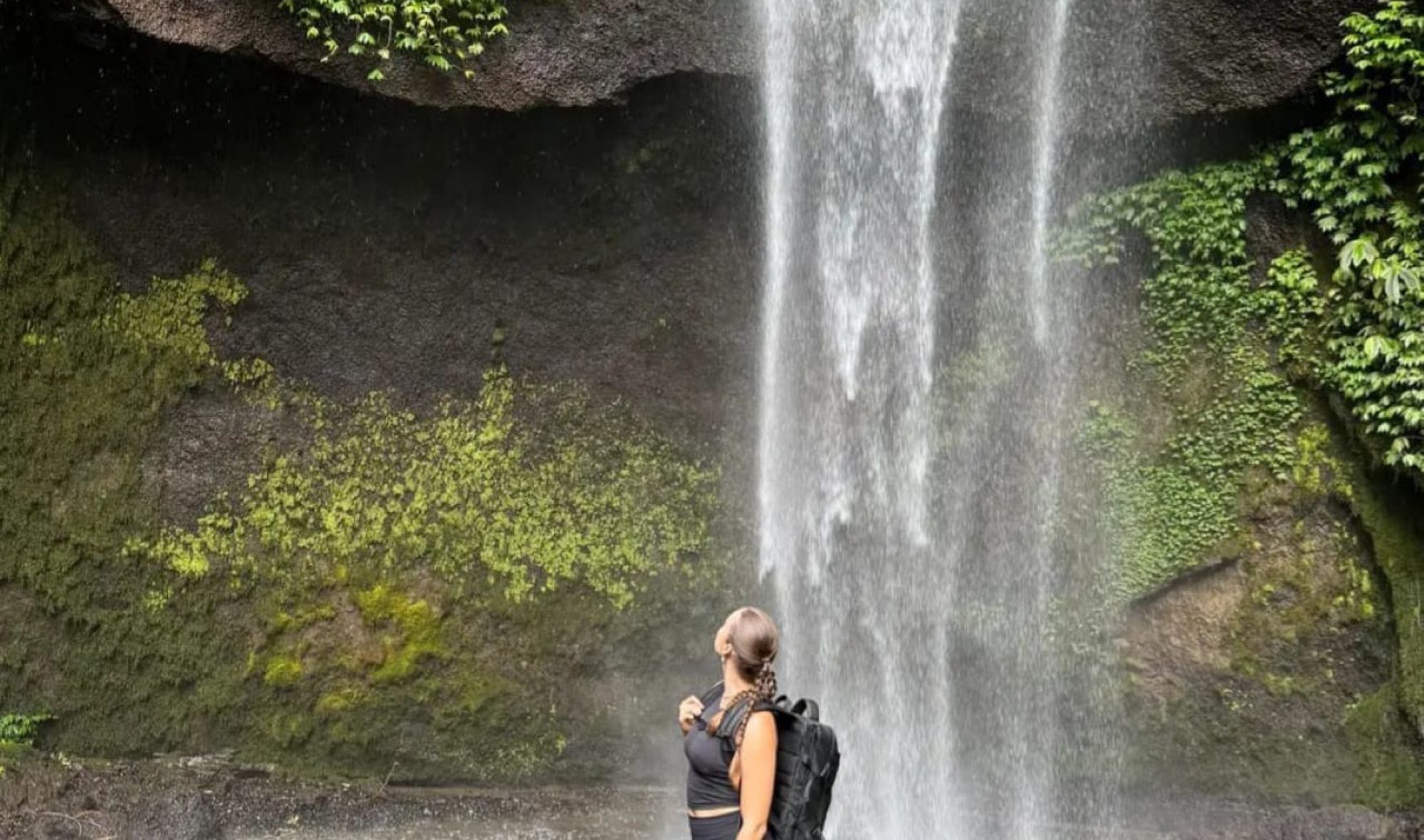 Woman wearing a backpack stands at the base of a tall waterfall, looking up as water cascades down a moss-covered cliff, with a small dog beside her.